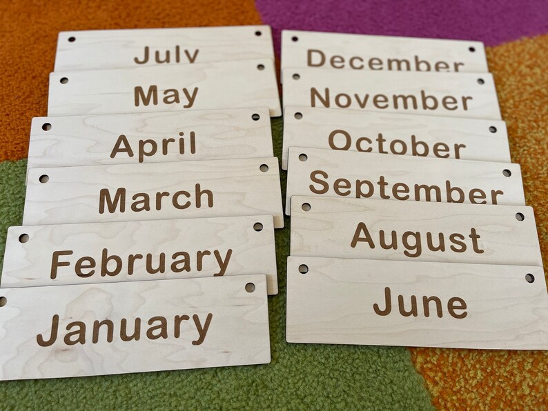 May include: Wooden month name plaques. Each plaque is rectangular with rounded corners and two small holes. The months are printed in a brown font. The months are: July, May, April, March, February, January, December, November, October, September, August, and June.