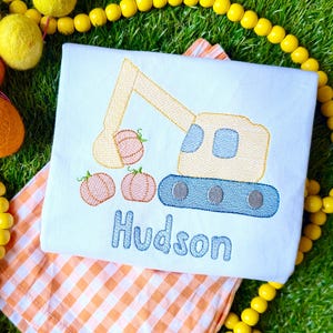 May include: White, embroidered shirt featuring a yellow and blue excavator design with three pumpkins. The name "Hudson" is embroidered in blue below the excavator. The shirt is on a checkered orange and white cloth, surrounded by yellow and orange decorative beads.