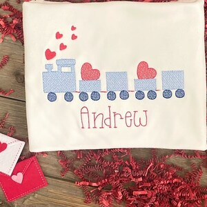 May include: A white onesie with a blue train carrying red hearts and the name "Andrew" embroidered on it. The onesie is surrounded by red paper shreds and a red heart with white polka dots.