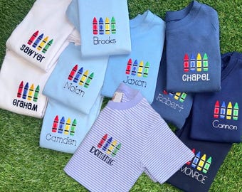 May include: A collection of short-sleeved shirts in white, light blue, dark blue, and blue-and-white stripes. Each shirt features an embroidered design of four crayons in red, blue, yellow, and green, with a unique name stitched below each.