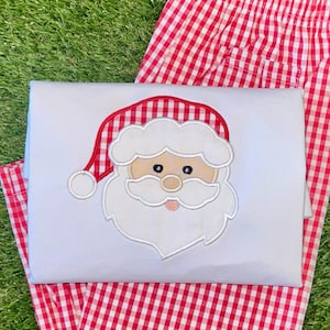 Puede incluir: Un mono blanco con una cara de Papá Noel bordada en rojo y blanco. El mono se combina con pantalones de cuadros rojos y blancos.