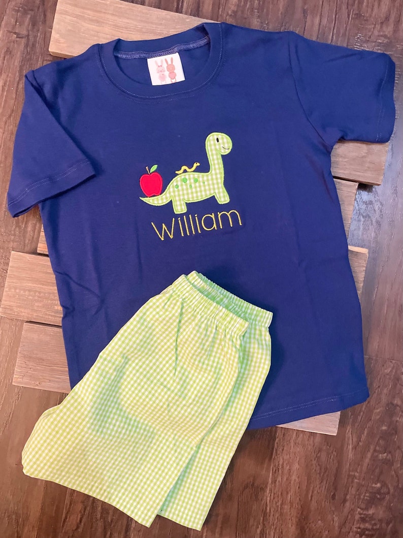 May include: A navy blue short-sleeved t-shirt with a green dinosaur and red apple applique. The shirt is personalized with the name "William". The shirt is paired with green and white gingham shorts.