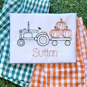 May include: A white t-shirt with an embroidered design of a green tractor pulling a wagon full of orange pumpkins. The name "Sutton" is embroidered below the tractor. The t-shirt is laying on top of a green and white gingham fabric and an orange and white gingham fabric.