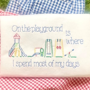 May include: White t-shirt with embroidered playground scene and the text "On the playground is where I spend most of my days" in blue thread. The playground includes a slide, swings, and a seesaw.
