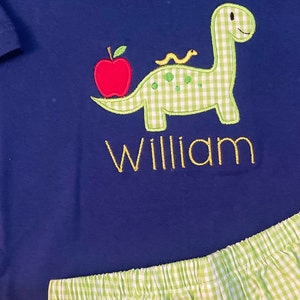 May include: A blue and green gingham dinosaur with a red apple and a yellow worm on its back. The dinosaur is embroidered on a blue shirt with the name "William" embroidered below it.