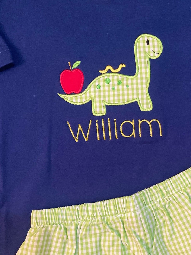 May include: A blue and green gingham dinosaur with a red apple and a yellow worm on its back. The dinosaur is embroidered on a blue shirt with the name "William" embroidered below it.