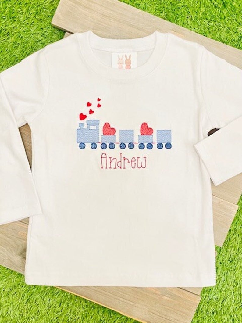 May include: White long-sleeve shirt with a blue and red embroidered train with hearts and the name "Andrew" on it.