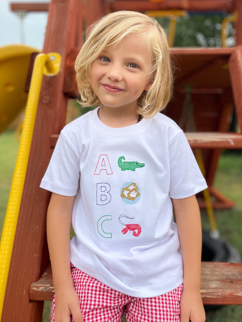 May include: A white t-shirt with the letters A, B, and C embroidered on the front. Each letter is paired with a different embroidered image: an alligator, a seashell, and a crawfish. The shirt is being worn by a young child.