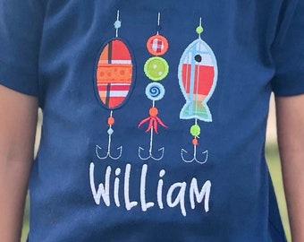 May include: A navy blue t-shirt with a colourful embroidered design of fishing lures and the name "William" in white.