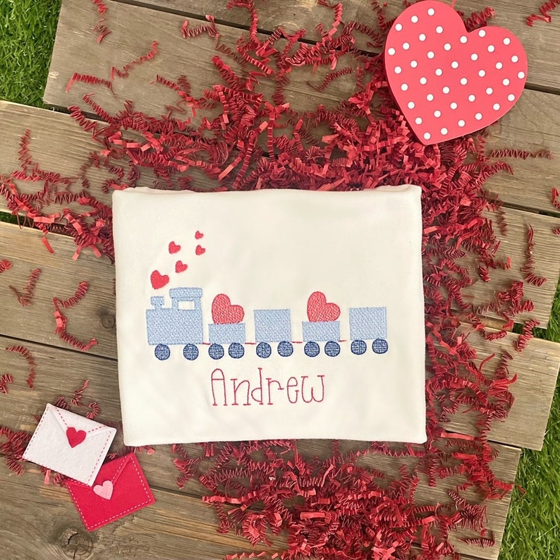 May include: A white onesie with a blue train carrying red hearts and the name "Andrew" embroidered on it. The onesie is surrounded by red paper shreds and a red heart with white polka dots.