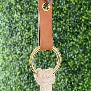 May include: A plant hanger featuring a tan leather strap with a brass ring and a knotted, off-white rope. The metal hook is silver. The background is a blurred green.