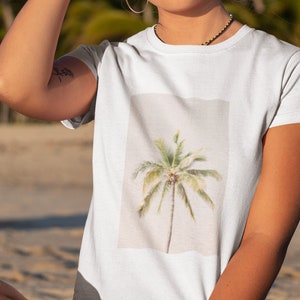 May include: White t-shirt featuring a watercolor-style palm tree design. The shirt is short-sleeved and has a crew neck. The background shows a beach with palm trees and a clear blue sky.