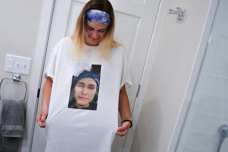Op de afbeelding: Wit T-shirt met een afgedrukte afbeelding van een persoon met een blauwe hoofdband. De persoon op de afbeelding op het shirt draagt ook een blauwe hoofdband. De persoon op de afbeelding glimlacht. Het shirt wordt omhoog gehouden.