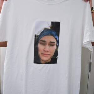 Op de afbeelding: Wit T-shirt met een rechthoekige afbeelding van een persoon met een blauwe hoofdband. De persoon op de afbeelding heeft een neutrale uitdrukking. Het T-shirt wordt door twee handen omhoog gehouden.