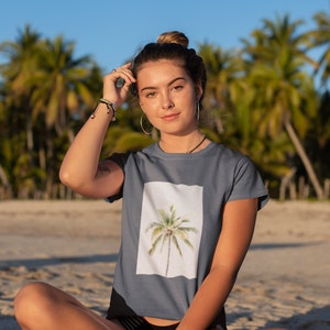 May include: A slate blue t-shirt featuring a watercolor image of a palm tree on a white background. The person is sitting on a beach with palm trees in the background.