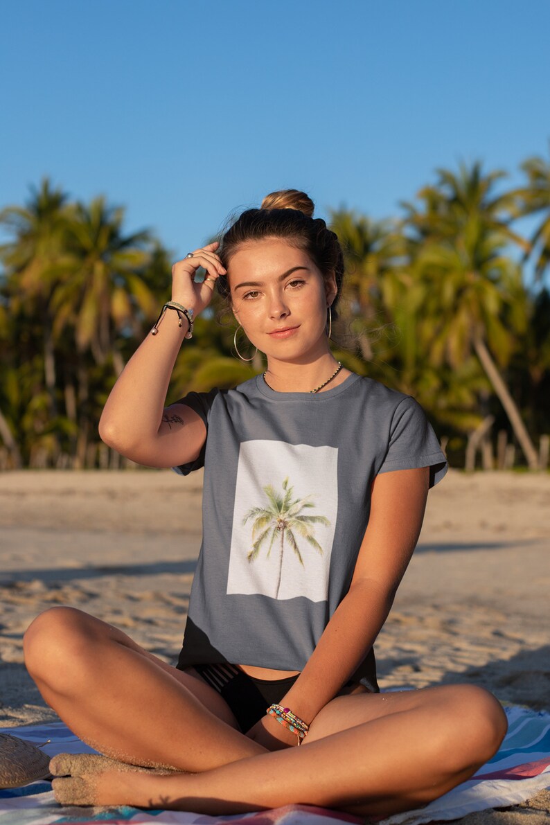 May include: A slate blue t-shirt featuring a watercolor image of a palm tree on a white background. The person is sitting on a beach with palm trees in the background.
