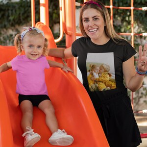 Op de afbeelding: Een jong kind in een roze t-shirt en zwarte shorts glijdt van een oranje glijbaan. Een volwassene draagt een zwart t-shirt met een fotoprint van twee kinderen. De achtergrond omvat een speeltuin en groen.