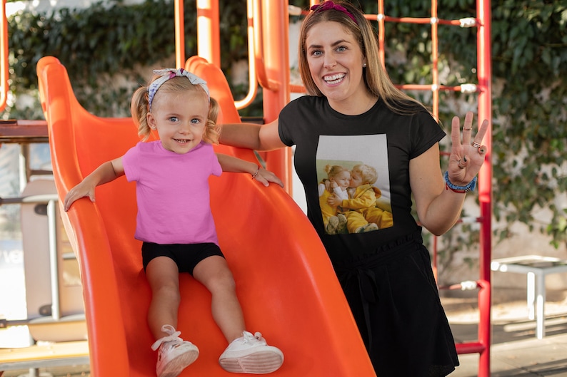 Op de afbeelding: Een jong kind in een roze t-shirt en zwarte shorts glijdt van een oranje glijbaan. Een volwassene draagt een zwart t-shirt met een fotoprint van twee kinderen. De achtergrond omvat een speeltuin en groen.