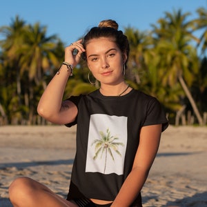 May include: Black t-shirt featuring a watercolor image of a palm tree on a white background. The model is sitting on a sandy beach with palm trees in the background. The shirt has short sleeves.