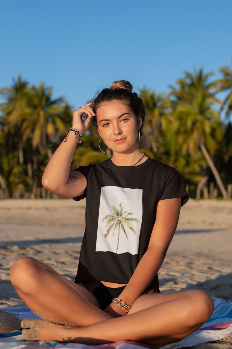 May include: Black t-shirt featuring a watercolor image of a palm tree on a white background. The model is sitting on a sandy beach with palm trees in the background. The shirt has short sleeves.