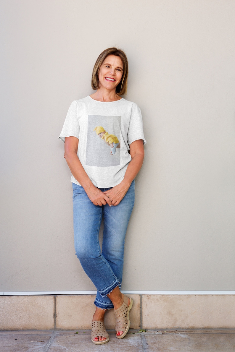 Op de afbeelding: Een vrouw draagt een lichtgrijs t-shirt met een rechthoekige afbeelding van drie figuren in gele hoeden en outfits. Ze draagt ook een blauwe spijkerbroek en bruine sandalen. De vrouw glimlacht en leunt tegen een lichtgrijze muur.