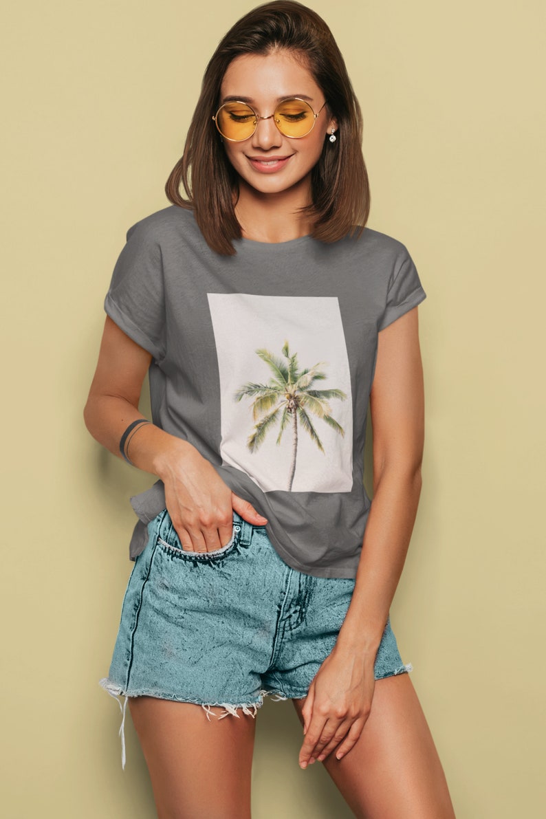 May include: Gray t-shirt featuring a rectangular image of a palm tree. The palm tree has a brown trunk and green fronds. The model is wearing blue denim shorts and yellow-tinted glasses.
