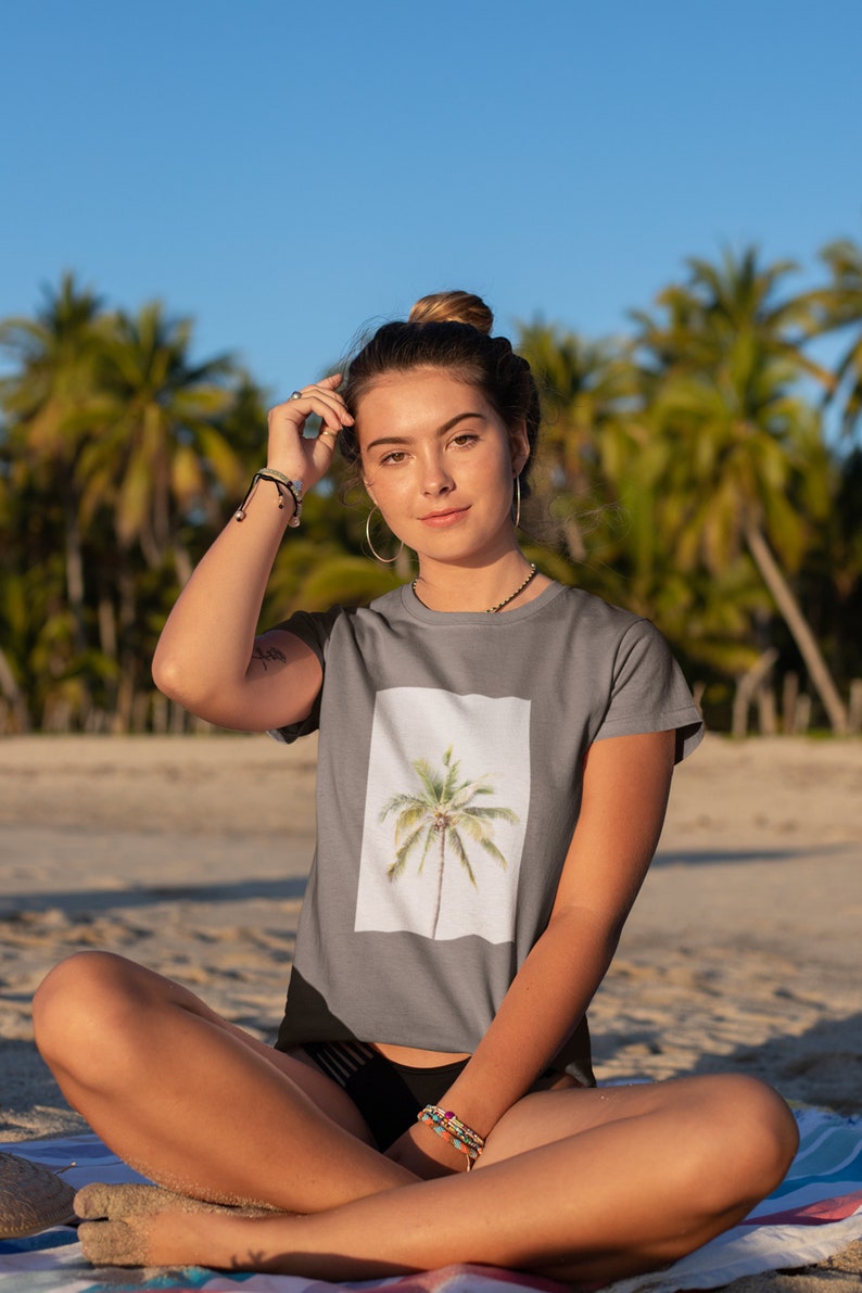May include: Gray t-shirt featuring a watercolor-style palm tree design within a white square. The shirt is short-sleeved and made of a soft material. The background shows a beach with palm trees.