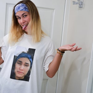 Op de afbeelding: Wit T-shirt met een afgedrukte afbeelding van een persoon met een blauwe hoofdband en een verdrietige uitdrukking. De persoon op de afbeelding draagt ook een blauwe hoofdband. De persoon op de afbeelding steekt zijn tong uit en draagt een blauwe hoofdband.