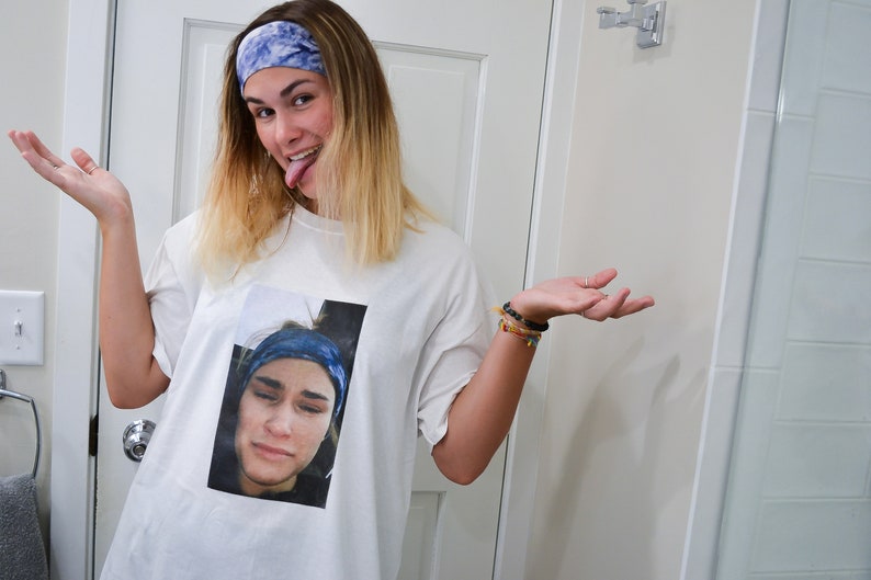 Op de afbeelding: Wit T-shirt met een afgedrukte afbeelding van een persoon met een blauwe hoofdband en een verdrietige uitdrukking. De persoon op de afbeelding draagt ook een blauwe hoofdband. De persoon op de afbeelding steekt zijn tong uit en draagt een blauwe hoofdband.
