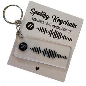 Peut inclure: Un porte-clés Spotify en acrylique transparent avec un anneau argenté. Le porte-clés présente un logo Spotify noir et un motif d'onde sonore. La carte derrière le porte-clés indique "Spotify Keychain" et "Sunflower - Post Malone, Swae Lee."