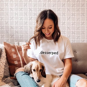 May include: A woman wearing a white t-shirt with the text ".desserped" printed on it, sitting on a couch with a small dog. The woman is wearing blue jeans and has brown hair. The couch is gray and has a brown and white throw pillow.