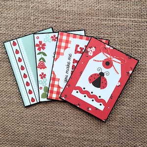 May include: Four decorative greeting cards with black borders. Designs include ladybugs, floral patterns, and gingham. One card features a ladybug tag with the text "you make me". The cards are arranged on a textured brown surface.