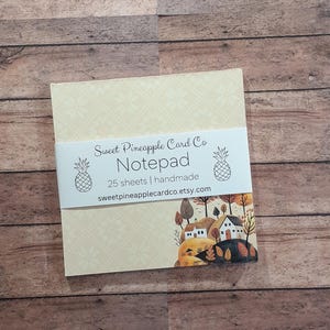 May include: A notepad from Sweet Pineapple Card Co. with a cream-colored background and a repeating floral pattern. The notepad has a white band with the company name, the word "Notepad", and a small illustration of a landscape with houses and trees.
