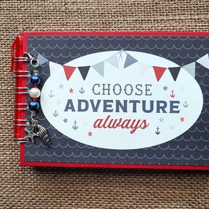 May include: A rectangular red notebook with a dark gray cover featuring a white oval center. The text CHOOSE ADVENTURE always is printed in the center, surrounded by bunting, stars, and anchors. A silver spiral binding holds blue and white beads.