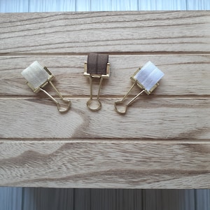 May include: Three metal binder clips with gold-toned arms and bodies. Each clip features a strip of fabric attached to the top edge: one in light beige, one in dark brown, and one in white. The clips are arranged in a horizontal row.
