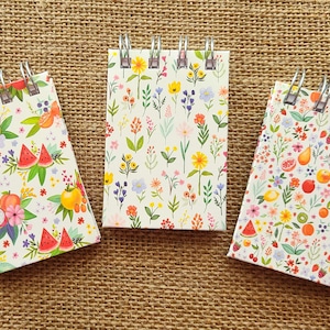 May include: Three spiral-bound notebooks with white covers featuring colorful patterns. The left notebook displays various fruits, the center notebook shows a variety of small wildflowers, and the right notebook features a dense pattern of mixed fruits and flowers.