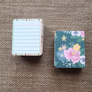 May include: Two small rectangular notepads. One notepad features a white interior with horizontal blue lines and a striped border. The other notepad displays a dark green cover decorated with a colorful floral pattern of pink, yellow, and blue flowers.