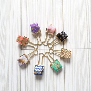 May include: A set of ten gold binder clips with different patterned fabric covers. The patterns include polka dots, arrows, geometric shapes, and a unicorn.