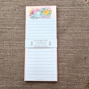 May include: A lined notepad with a floral design at the top, featuring a teapot and roses. The notepad is light pink with horizontal lines for writing. A white band with text and pineapple graphics wraps around the notepad.