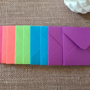 May include: A collection of colorful, small envelopes in shades of pink, orange, green, blue, and purple. The envelopes are arranged in a row on a textured, neutral-toned surface. A white decorative object is in the background.