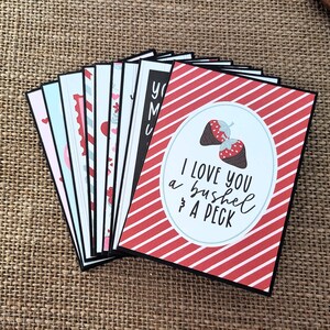 May include: A stack of greeting cards with various designs. The front card has a red and white striped pattern with the text "I love you a bushel & a peck" and a strawberry graphic. Other cards have hearts and other designs.