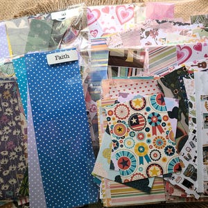 May include: A collection of patterned paper sheets in various colors and designs. Includes blue polka dots, floral, striped, and patriotic patterns. Some sheets are labeled with the words "Faith" and "ai". Ideal for scrapbooking, card making, and other paper crafts.