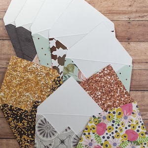 May include: A collection of decorative envelopes with various patterned designs. The envelopes have white interiors and feature patterns such as gold glitter, floral, and geometric shapes. The envelopes are arranged on a wooden surface.