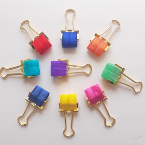 May include: Eight gold binder clips with colorful elastic bands. The bands are red, blue, turquoise, purple, green, pink, yellow, and orange.
