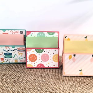 May include: A collection of decorative desk organizers with colorful patterns. Designs include sewing notions, smiley faces, and lemons. Each organizer has a rectangular base and a slot for holding papers or notes.