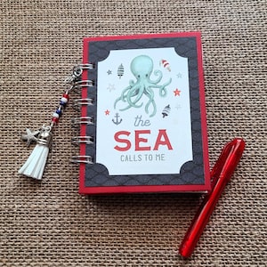 May include: A spiral-bound notebook with a red cover and a central white label featuring a light blue octopus, small nautical icons, and the text "the SEA CALLS TO ME". A silver charm with a white tassel and red, white, and blue beads is attached.