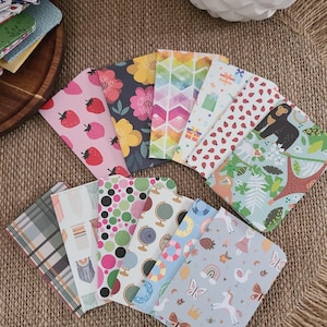 May include: A collection of 12 small, colorful notebooks with various patterns, including strawberries, flowers, geometric shapes, and animals. The notebooks are arranged in a fan shape on a brown burlap surface.