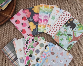 May include: A collection of 12 small, colorful notebooks with various patterns, including strawberries, flowers, geometric shapes, and animals. The notebooks are arranged in a fan shape on a brown burlap surface.