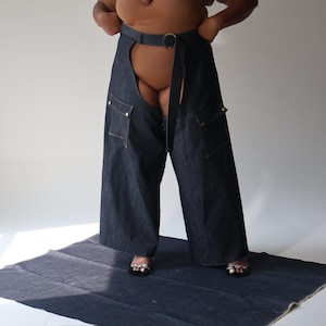 May include: Dark wash denim chaps with large pockets and a belt with a gold buckle. The chaps are open in the back and worn over a nude bodysuit. The model is standing on a dark blue fabric.
