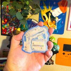 May include: A blue and white sticker of a vintage Sony Handycam camcorder with glitter accents. The sticker has a white star and the text "HANDYCAM" in rainbow colors.